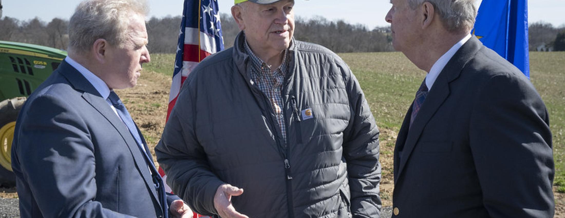 Three men with flag and farm field in background.